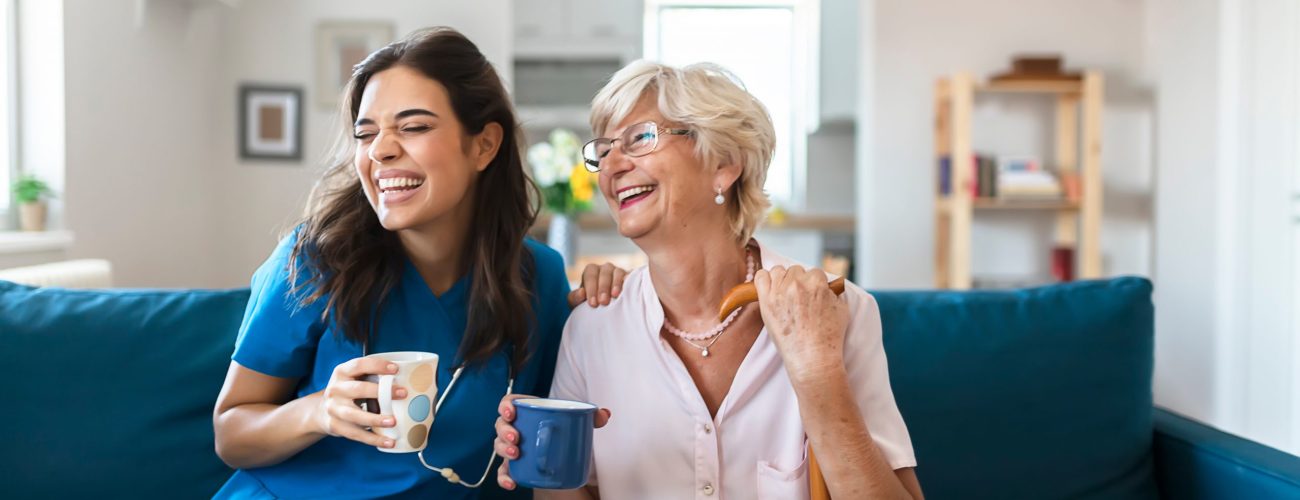 Photo of young caregiver laughing with senior resident in living room during home visit.